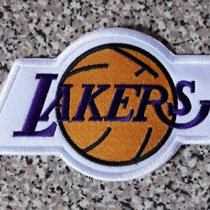 May include: An embroidered patch featuring the word LAKERS in purple text across a yellow basketball graphic. The design is set against a white background with a white border, creating a distinct shape that highlights the team logo branding.