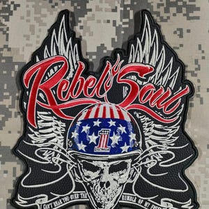 Puede incluir: Un parche negro con las palabras "Rebel's Soul" en letras rojas, con una calavera que lleva un casco con estrellas y rayas. Alas blancas y el texto "I can't hear you over the rumble of my freedom" completan el diseño.
