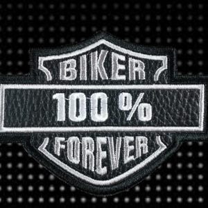 Op de afbeelding: Zwart-witte biker patch met de tekst "BIKER 100% FOREVER". De patch heeft een schildvorm, witte stiksels en een getextureerde, lederachtige uitstraling.