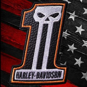 Puede incluir: Un parche bordado en blanco y negro con forma de número uno, que presenta un diseño de calavera integrado en la parte superior del dígito y el texto Harley-Davidson en letras negras en la base, delineado con un borde naranja fino.