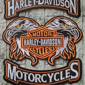 Puede incluir: Un conjunto de tres parches bordados con el logotipo de Harley-Davidson en blanco y naranja sobre un fondo negro. Las bandas superior e inferior muestran el nombre de la marca y la palabra Motorcycles, mientras que la pieza central muestra dos águilas.
