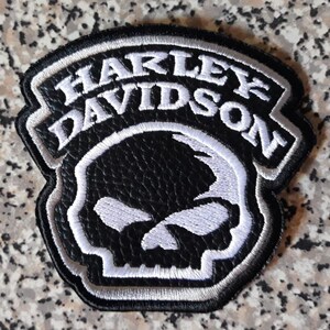 Puede incluir: Un parche bordado en blanco y negro que presenta el texto Harley Davidson en una fuente curva sobre un gráfico de calavera estilizada. El diseño está delineado con un borde blanco grueso y tiene una superficie texturizada como cuero.