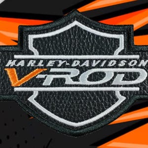 Puede incluir: Parche Harley-Davidson V-Rod en blanco y negro con apariencia de cuero texturizado. El parche presenta el logotipo de Harley-Davidson y el texto V-Rod en blanco, con una "V" naranja. El fondo es negro y naranja.