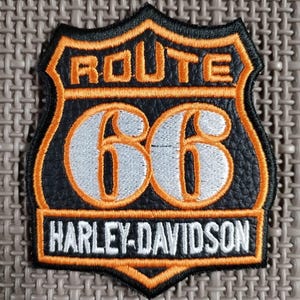 Puede incluir: Un parche negro y naranja de Route 66 Harley-Davidson. El parche presenta las palabras "ROUTE 66" en naranja, el número "66" en plata y "HARLEY-DAVIDSON" en blanco. El parche tiene un borde naranja.