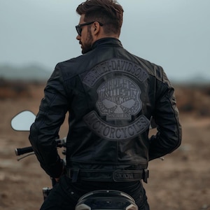 May include: Black leather motorcycle jacket with the Harley-Davidson logo on the back. The jacket features a skull design within the logo and is worn by a person on a motorcycle. The jacket has a classic biker style.