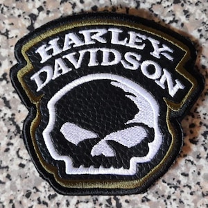 Puede incluir: Un parche bordado en blanco y negro que presenta el texto Harley Davidson en letras blancas arqueadas sobre un gráfico de calavera blanca estilizada. El parche tiene un fondo con textura de cuero negro y un borde fino de color dorado.