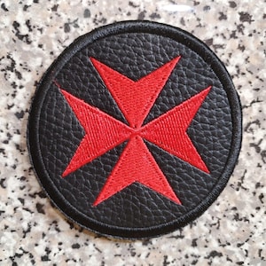 May include: A round, black patch with a red Maltese cross design. The cross is prominently featured in the center, with a textured black background and a stitched border. The patch is likely for clothing or accessories.