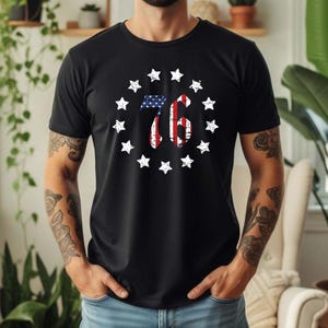 Puede incluir: Camiseta negra con un diseño circular. El diseño presenta el número "76" en rojo y blanco, relleno con un patrón de la bandera estadounidense, rodeado de estrellas blancas.