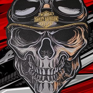 Puede incluir: Un parche bordado en negro, blanco y dorado que muestra un cráneo humano con gafas protectoras. El texto Harley-Davidson Motor Cycles está bordado en hilo dorado sobre la frente. El diseño incluye sombreados y líneas de contorno detalladas.