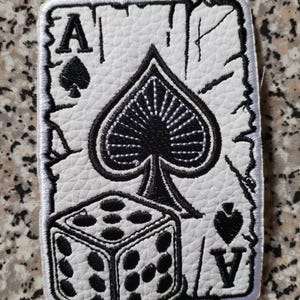 Puede incluir: Un parche blanco con un diseño de carta de As de Picas y un dado en negro. El parche tiene un aspecto envejecido y texturizado con costuras negras. El diseño incluye la letra "A" y los símbolos de picas y corazones.