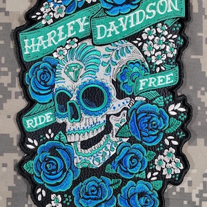 Puede incluir: Parche bordado con un diseño de calavera de azúcar en turquesa y blanco, un diamante y rosas azules y flores blancas. El parche incluye las palabras "HARLEY DAVIDSON" y "RIDE FREE" en pancartas verdes.