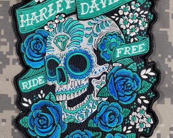 May include: Embroidered patch featuring a sugar skull design with turquoise and white detailing, a diamond, and surrounded by blue roses and white flowers. The patch includes the words "HARLEY DAVIDSON" and "RIDE FREE" on green banners.