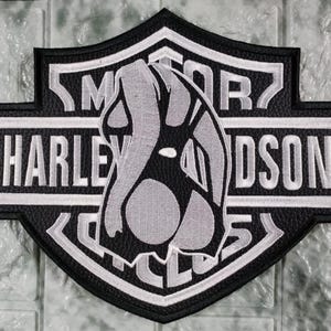 Op de afbeelding: Een zwart-witte geborduurde patch in de vorm van een schild met de tekst Harley-Davidson. De patch toont een centrale afbeelding van een gestileerd figuur in een zittende houding, uitgevoerd in grijze en zwarte draad.
