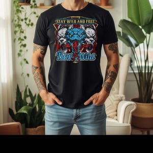 Puede incluir: Camiseta negra con un diseño gráfico de una serpiente azul y dos cabezas de lobo blancas. El texto "STAY WILD AND FREE" está encima de la serpiente y "Born Wild" debajo. La camiseta la lleva una persona con vaqueros azules.