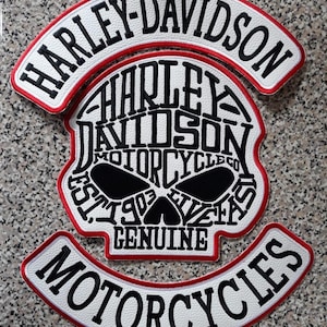 May include: Embroidered Harley-Davidson motorcycle patch set. The set includes a skull-shaped patch with the words "Harley-Davidson Motorcycle Co. Est. 1903 Live Fast Genuine" and two arched patches with the words "Harley-Davidson" and "Motorcycles". The patches have a white background with black text and red trim.