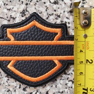 Op de afbeelding: Zwart en oranje geborduurde patch met een schildontwerp. De patch is ongeveer 6,35 cm breed. De patch heeft een getextureerd uiterlijk en is waarschijnlijk gemaakt van leer of een vergelijkbaar materiaal.