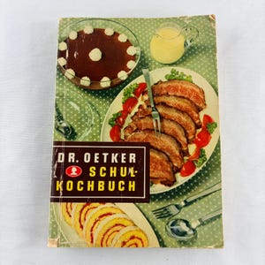 May include: A vintage Dr. Oetker Schulkochbuch cookbook with a green star-patterned cover. The cover features a chocolate dessert, a pitcher of yellow sauce, sliced roast meat with tomatoes, and a sliced cake roll, with silver cutlery.
