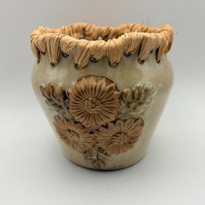 May include: A ceramic pot with a glossy beige finish featuring a raised relief of three brown sunflowers and leaves on the front. The rim is decorated with a woven natural raffia trim threaded through small holes around the top edge of the vessel.