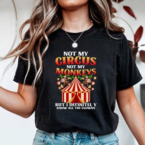 以下が含まれることがあります： サーカスをテーマにしたグラフィックがプリントされた黒いTシャツ。「NOT MY CIRCUS, NOT MY MONKEYS」の文字がオレンジと赤でデザインされ、サーカスのテントと猿が描かれています。「BUT I DEFINITELY KNOW ALL THE CLOWNS」のフレーズも含まれています。