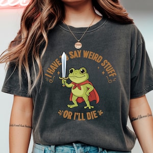 Puede incluir: Camiseta gris oscuro con un dibujo de una rana de dibujos animados con una capa roja y una espada. El texto dice "I HAVE TO SAY WEIRD STUFF OR I'LL DIE". La rana es verde con ojos grandes.