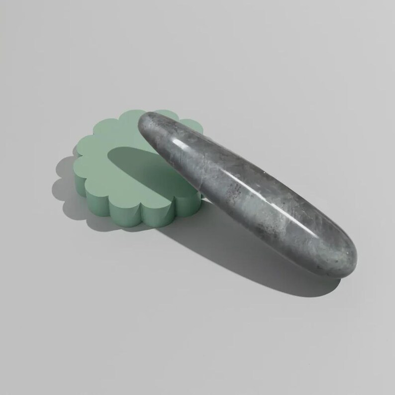 May include: A grey, elongated stone object with a smooth surface rests on a light green, scalloped platform. The stone has a marbled appearance. The background is a solid, light grey.