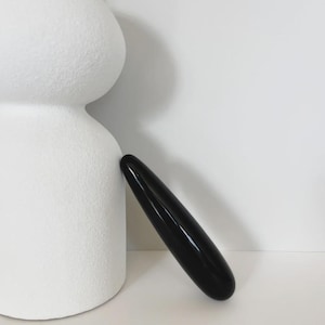 May include: A smooth, elongated, black cylindrical object with rounded ends is positioned diagonally against the side of a large, textured, matte white sculptural form that features two stacked, rounded, bulbous sections.