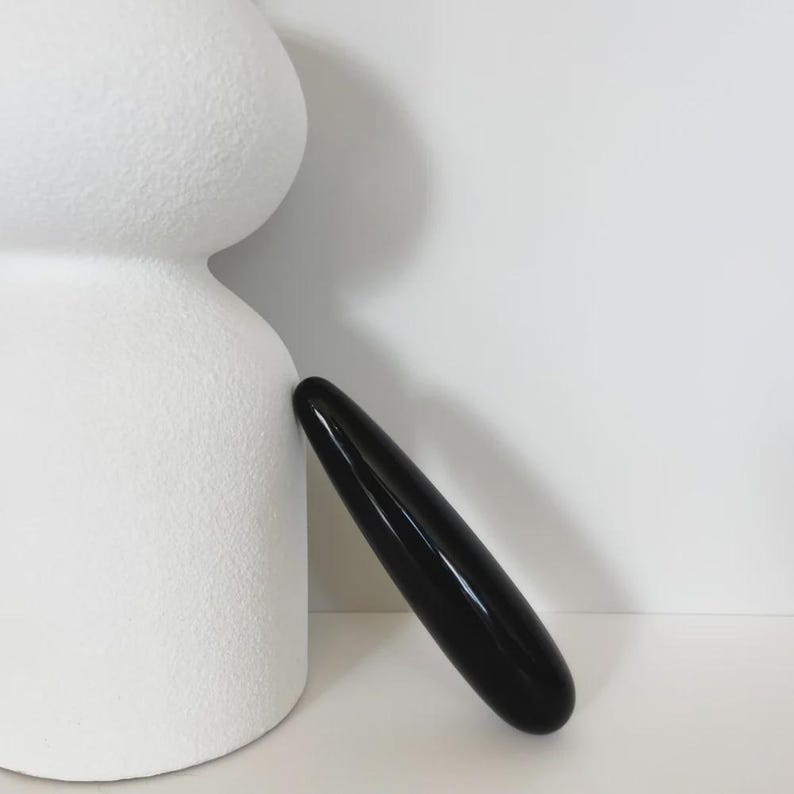 May include: A smooth, elongated, black cylindrical object with rounded ends is positioned diagonally against the side of a large, textured, matte white sculptural form that features two stacked, rounded, bulbous sections.