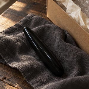 May include: A smooth, cylindrical, black stone massage wand with a polished, reflective surface. The object is elongated with rounded ends, featuring a solid, opaque black color throughout its entire length and a uniform, tapered shape.