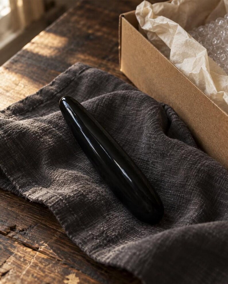 May include: A smooth, cylindrical, black stone massage wand with a polished, reflective surface. The object is elongated with rounded ends, featuring a solid, opaque black color throughout its entire length and a uniform, tapered shape.