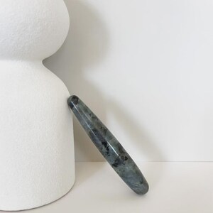 May include: A polished, elongated gemstone wand with a rounded tip. The wand is a mix of grey, blue, and black hues, with a glossy finish. It is leaning against a white, abstract ceramic sculpture.