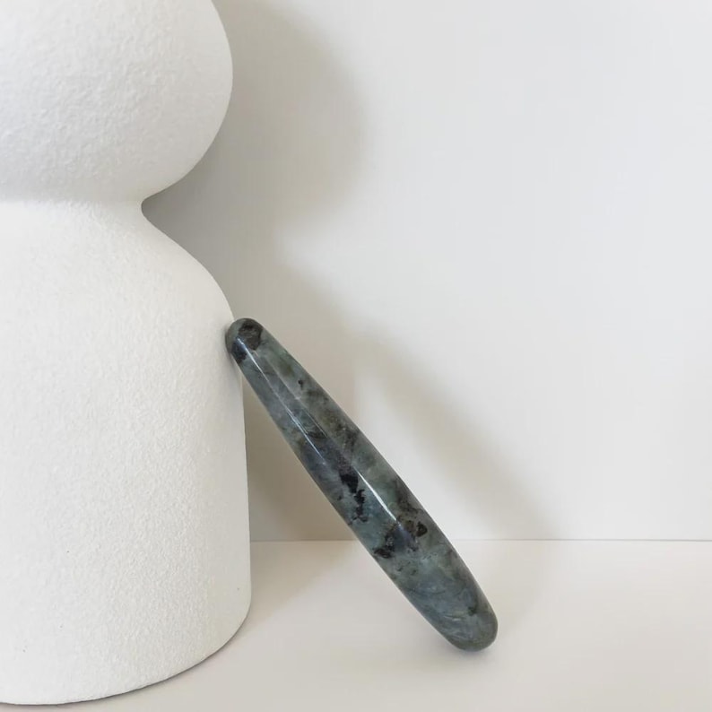 May include: A polished, elongated gemstone wand with a rounded tip. The wand is a mix of grey, blue, and black hues, with a glossy finish. It is leaning against a white, abstract ceramic sculpture.