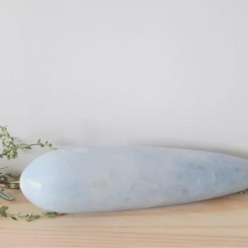May include: A pale blue, elongated stone with a smooth, polished surface. The stone is oval-shaped, wider at one end and tapering to a point. It rests on a light-colored wooden surface, with sprigs of greenery visible on the left side.