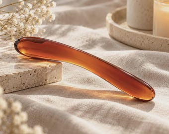 May include: A smooth, curved, amber-colored glass wand with a translucent finish. The object features a rounded tip at one end and a slightly tapered, rounded end at the other, showcasing a polished, reflective surface that catches the light.