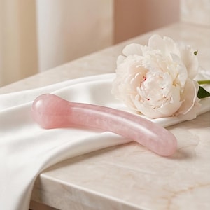 Puede incluir: Una herramienta de masaje facial de cuarzo rosa de color rosa, con un dise&ntilde;o curvo. La herramienta descansa sobre una tela de sat&eacute;n blanco junto a una gran flor de peon&iacute;a blanca. El fondo es de color neutro.