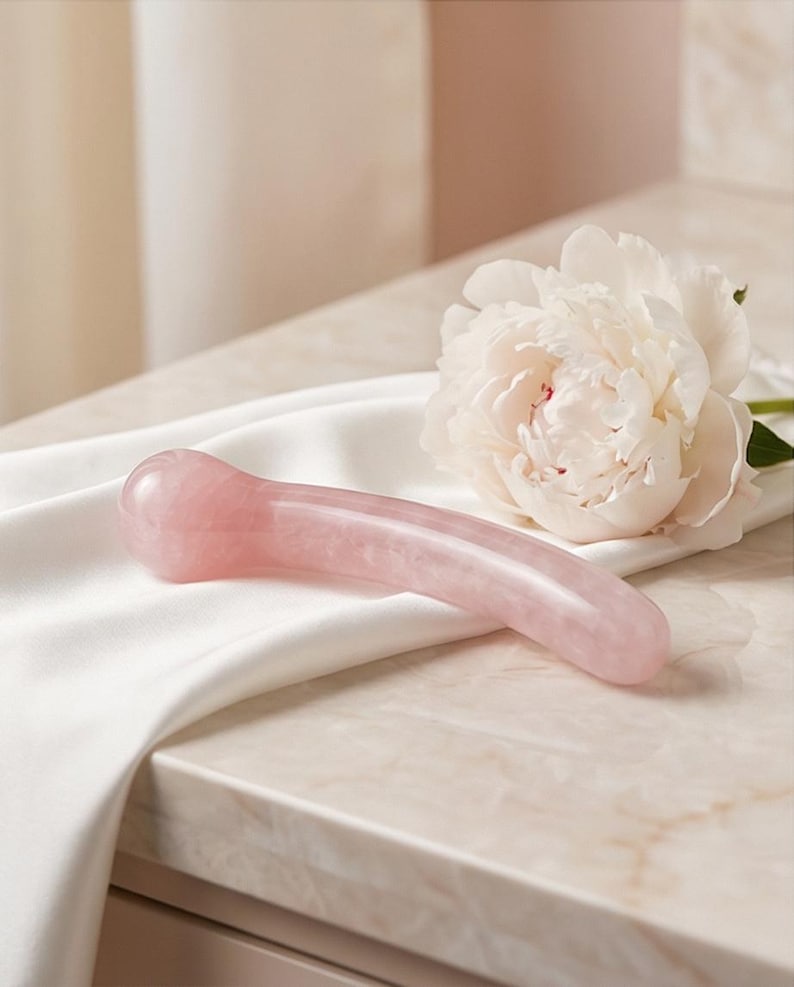 Puede incluir: Una herramienta de masaje facial de cuarzo rosa de color rosa, con un dise&ntilde;o curvo. La herramienta descansa sobre una tela de sat&eacute;n blanco junto a una gran flor de peon&iacute;a blanca. El fondo es de color neutro.