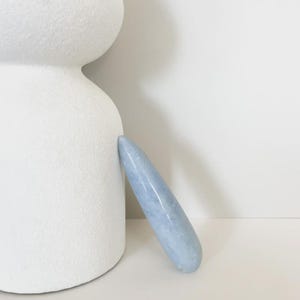 May include: A light blue crystal wand leans against a white, textured, rounded sculpture. The wand is smooth and elongated, with a tapered end. The background is a plain white surface.