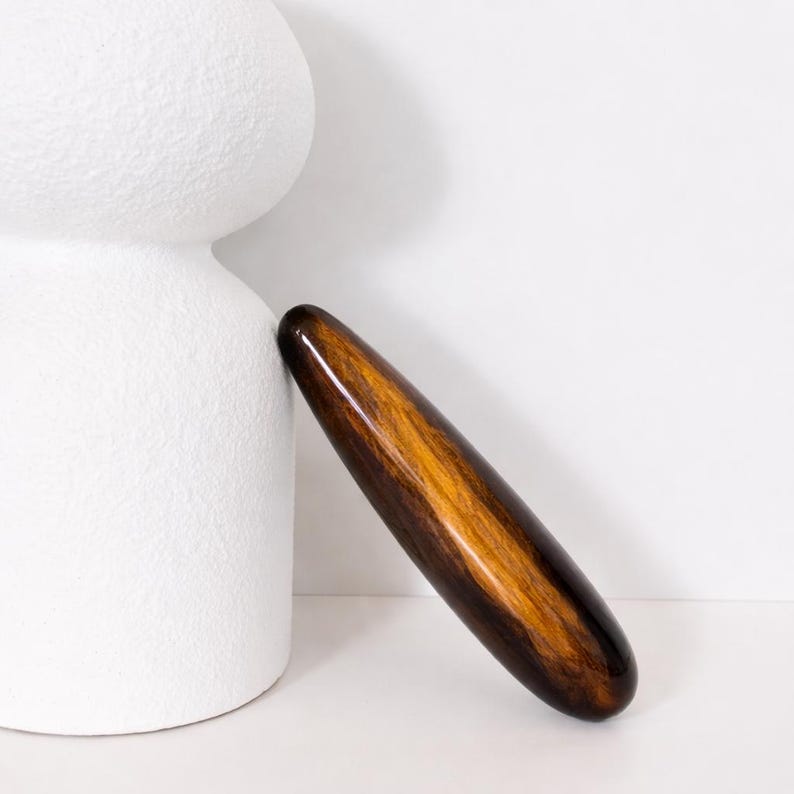 May include: A polished wooden massage tool with a dark brown and golden-brown grain pattern. The smooth, cylindrical shape is leaning against a textured white ceramic object, set against a white background. The tool is approximately 20 cm long.