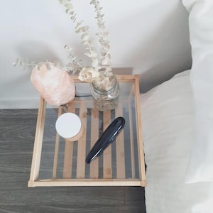 May include: A square wooden side table with a glass top holding a pink salt lamp, a clear glass jar containing dried eucalyptus stems, a small round container with a white lid, and a smooth, elongated black object with a curved silhouette.
