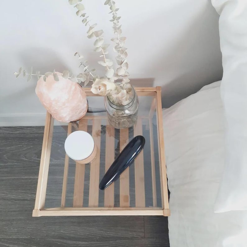 May include: A square wooden side table with a glass top holding a pink salt lamp, a clear glass jar containing dried eucalyptus stems, a small round container with a white lid, and a smooth, elongated black object with a curved silhouette.