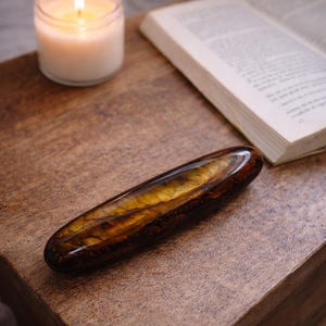 May include: A polished, elongated gemstone with brown and gold tones, placed on a wooden surface. A lit candle in a glass jar and an open book are also on the table, creating a cosy atmosphere.
