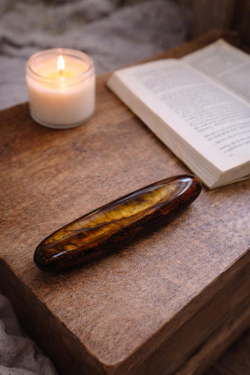May include: A polished, elongated gemstone with brown and gold tones, placed on a wooden surface. A lit candle in a glass jar and an open book are also on the table, creating a cosy atmosphere.