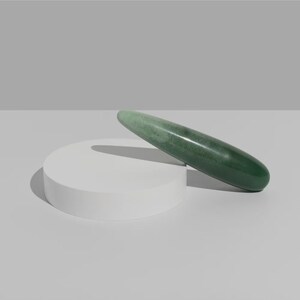 Op de afbeelding: Een gladde, groene jade massagetool ligt op een wit, cirkelvormig platform. De jade tool is langwerpig en cilindrisch, met een afgeronde punt. De achtergrond is neutraal grijs, wat de natuurlijke kleur en textuur van de jade benadrukt. Ongeveer 15 cm lang.