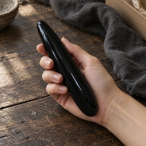 May include: A smooth, polished, cylindrical black stone massage wand with rounded ends. The object has a glossy, reflective surface and a solid, uniform dark color throughout its elongated, tapered shape, designed for handheld use.