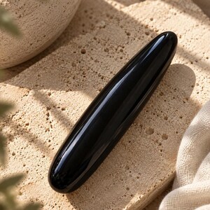 May include: A smooth, cylindrical, black stone wand with a polished, reflective surface. The object is elongated and tapers slightly at both rounded ends, featuring a solid, opaque black color throughout its entire length.