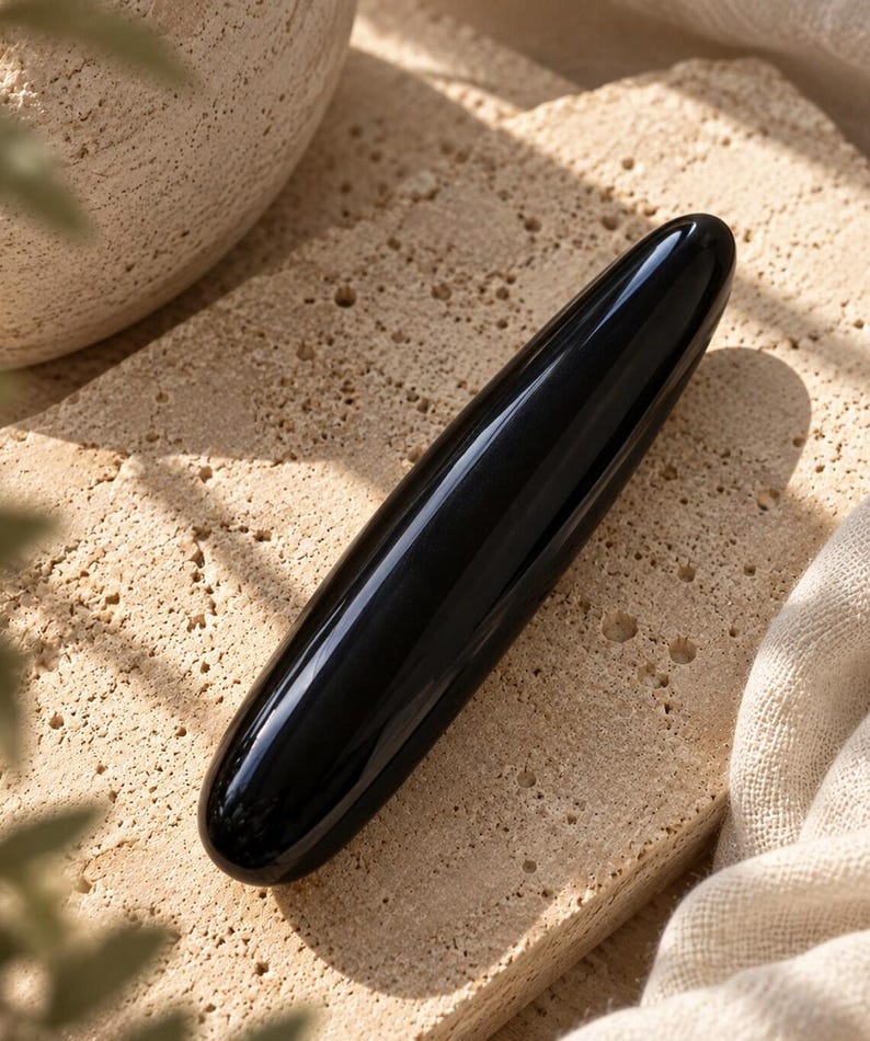 May include: A smooth, cylindrical, black stone wand with a polished, reflective surface. The object is elongated and tapers slightly at both rounded ends, featuring a solid, opaque black color throughout its entire length.