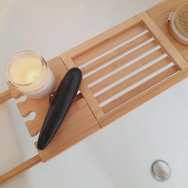 May include: A bamboo bathtub tray featuring a slatted surface, a lit candle in a glass jar, a smooth black elongated object, and a round wooden bath brush. The tray is expandable and includes a cutout section for holding items securely.