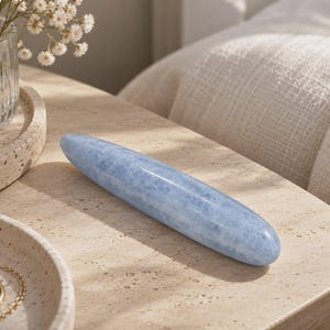 May include: A smooth, elongated, cylindrical stone object with a polished finish and a light blue color featuring subtle white and grey mineral inclusions throughout its solid, tapered, and rounded form.