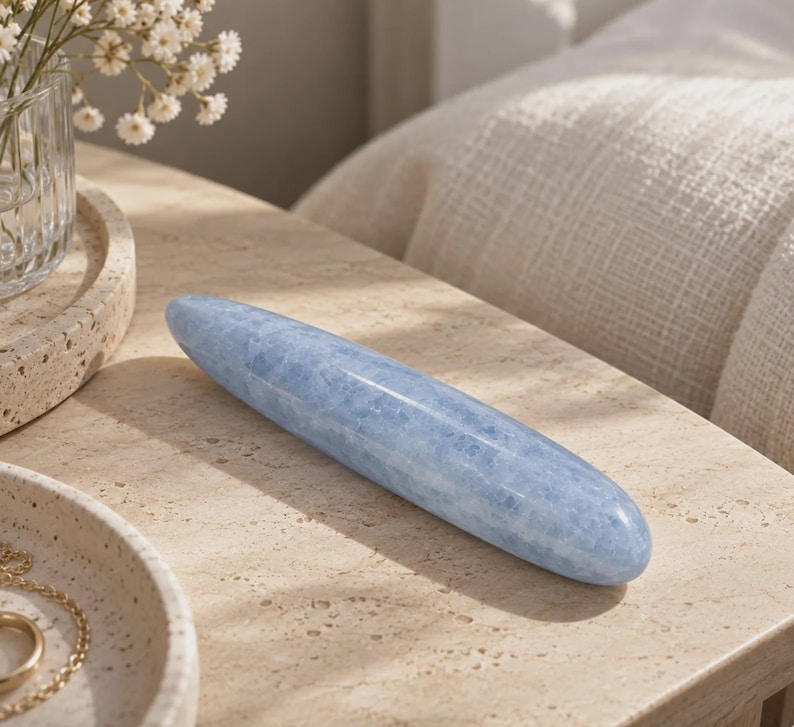 May include: A smooth, elongated, cylindrical stone object with a polished finish and a light blue color featuring subtle white and grey mineral inclusions throughout its solid, tapered, and rounded form.