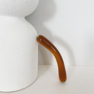 Könnte beinhalten: Eine weiße Keramik-Teekanne mit strukturierter Oberfläche und einem gebogenen, bernsteinfarbenen Griff. Der Griff ist an der Seite der Teekanne befestigt und schafft ein modernes und minimalistisches Design. Die Teekanne steht vor einem weißen Hintergrund.