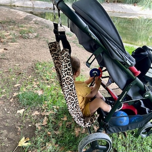 Peut inclure: Un bébé en tenue jaune est assis dans un hamac pour bébé à imprimé léopard, attaché à une poussette noire et grise. La poussette a un cadre noir et un panier de rangement avec des jouets bleus. L'arrière-plan montre un étang et de la verdure.